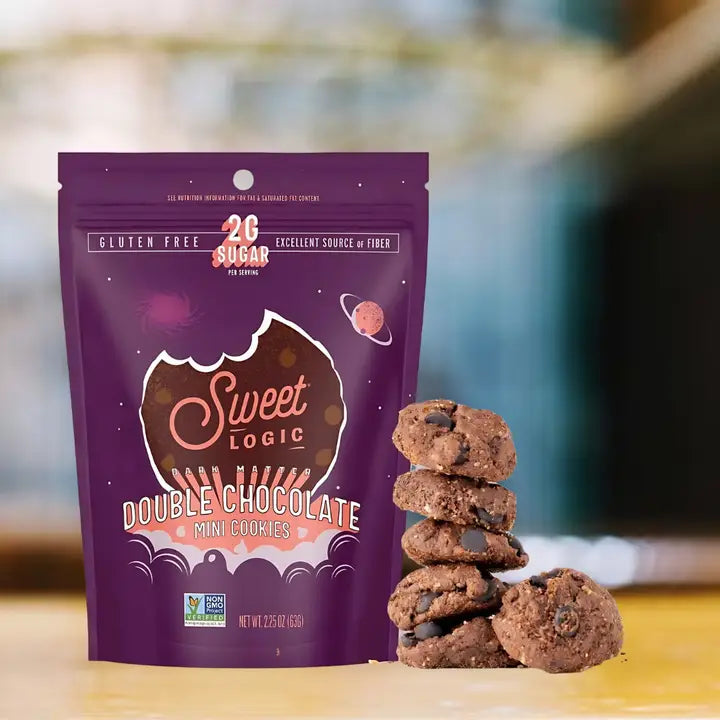 A purple stand-up pouch of Sweet Logic Double Chocolate Mini Cookies stands on a wooden surface with a stack of chocolate chip cookies beside it. The package shows the Sweet Logic brand, gluten free, 2C Sugar, and excellent source of fiber claims, with a softly blurred indoor background.
