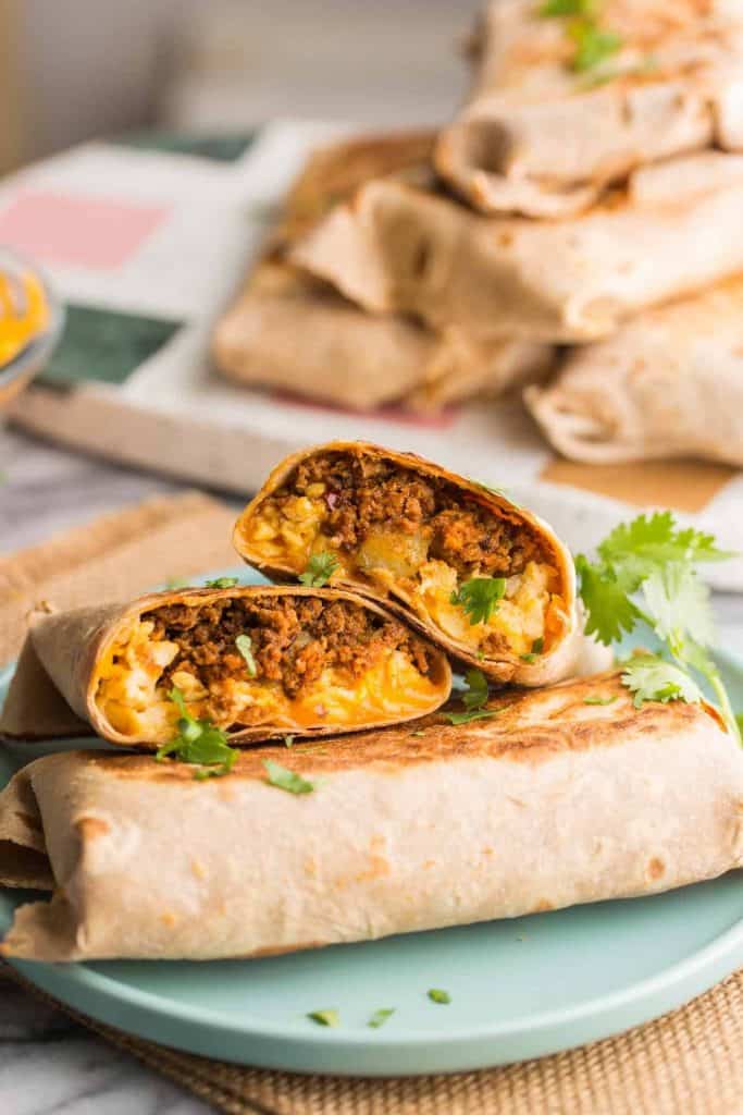A close-up food photo of several burritos on a light blue plate, with one burrito cut in half to show a filling of eggs, cheese, and ground chicken chorizo. Fresh cilantro is sprinkled on top, and more burritos are stacked in the blurred background on a countertop.