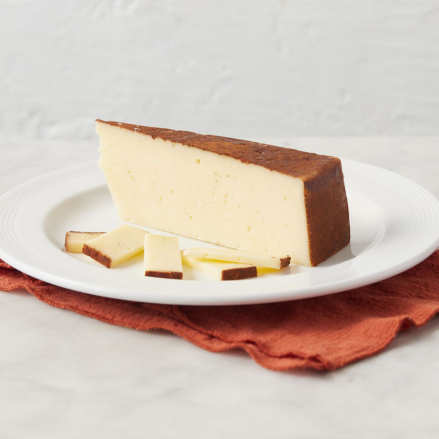A wedge of creamy off-white cheese with a dark brown rind, served on a white plate with small slices on top, resting on a rust-colored cloth against a light textured background.