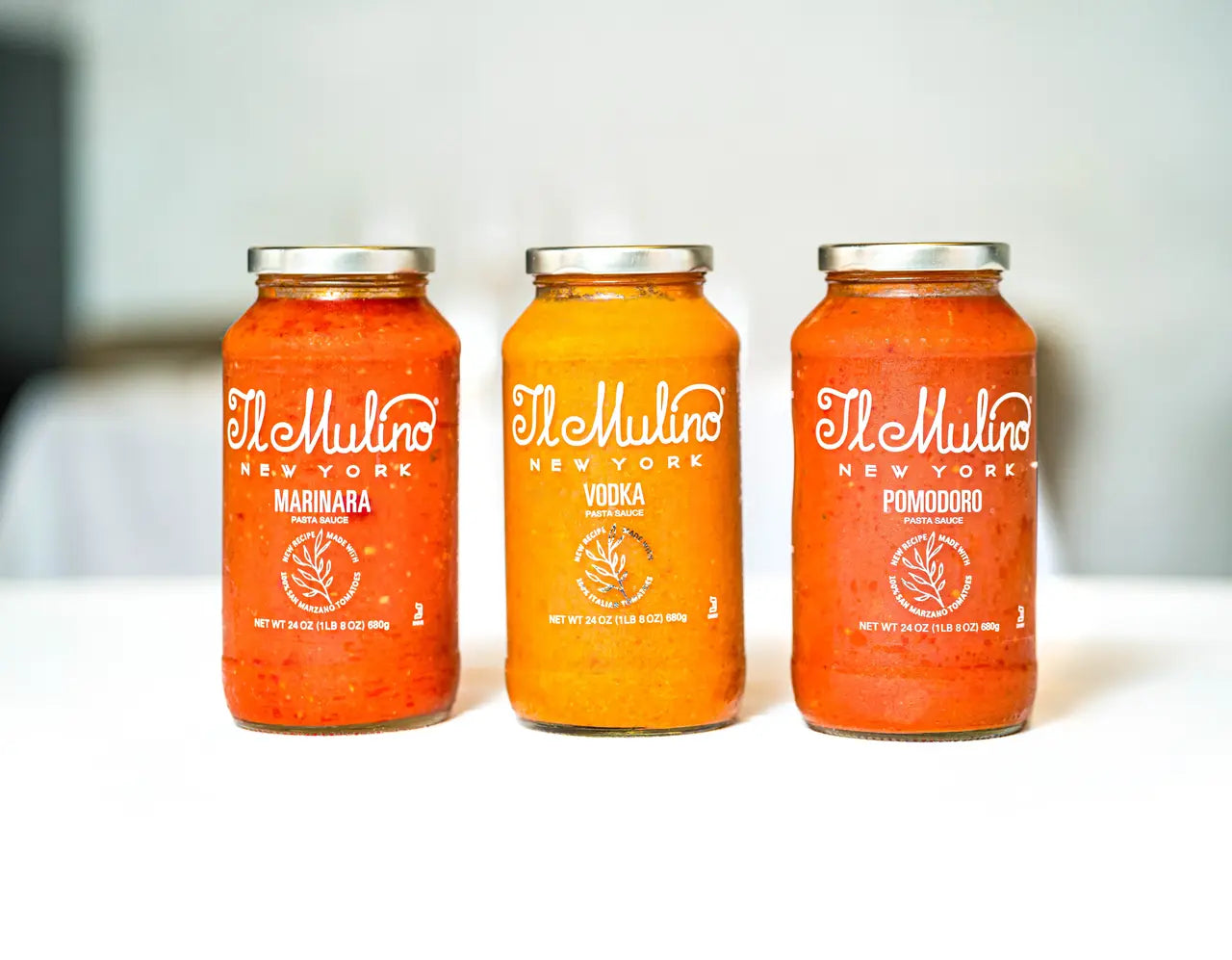 Three glass jars of The Mulino New York marinara sauce in different flavors (Marinara, Vodka, and Pomodoro) lined up on a white surface against a blurred light gray background.
