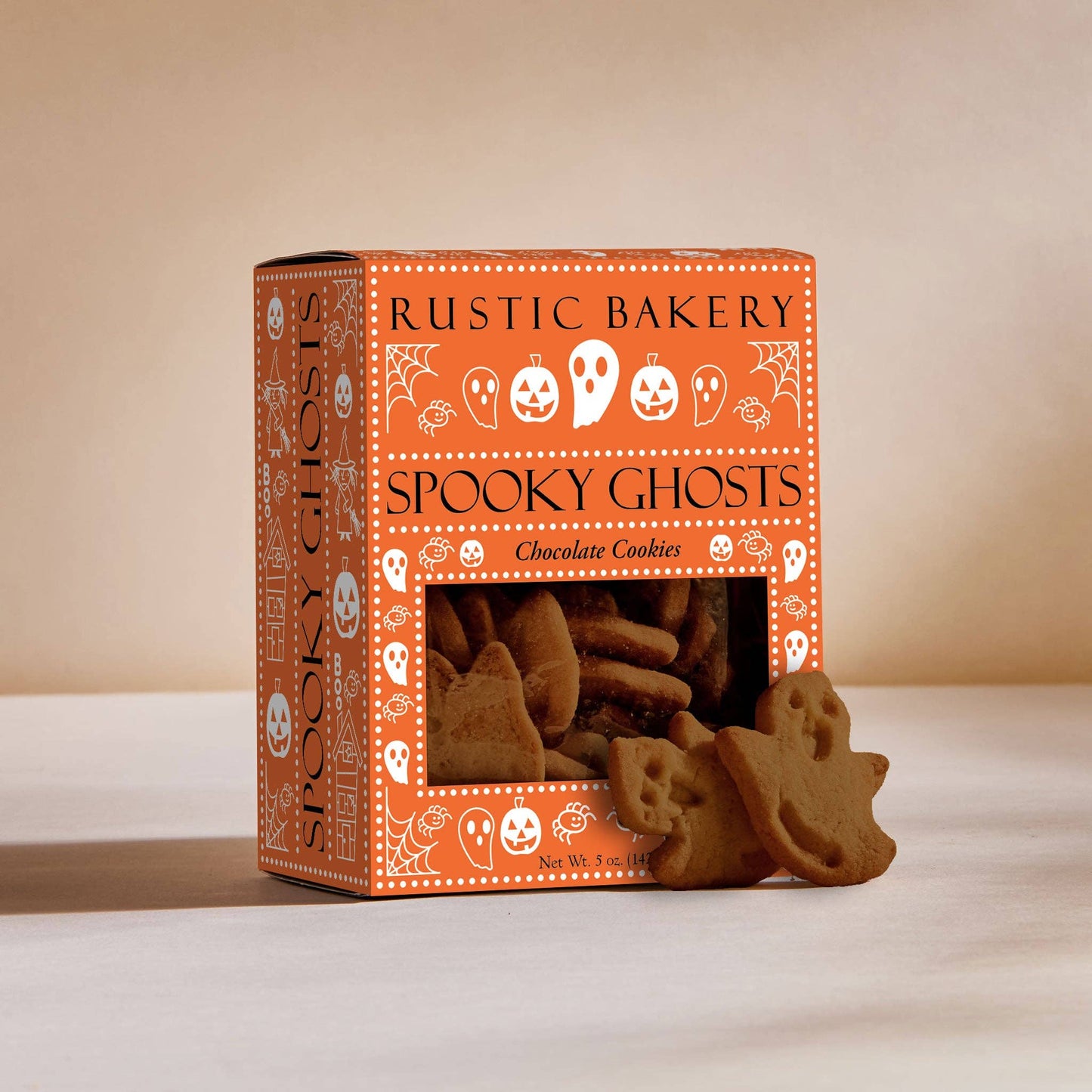 A box of Rustic Bakery Spooky Ghosts chocolate cookies on a light beige surface with a matching background. The orange packaging features white Halloween-themed illustrations, and several ghost-shaped cookies are displayed in front of the window.
