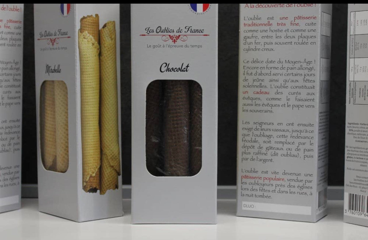 Three white bakery boxes displayed side-by-side on a white surface, showing different French pastries: a chocolate pastry with a window, a waffle cone pastry, and a dark chocolate bar inside.