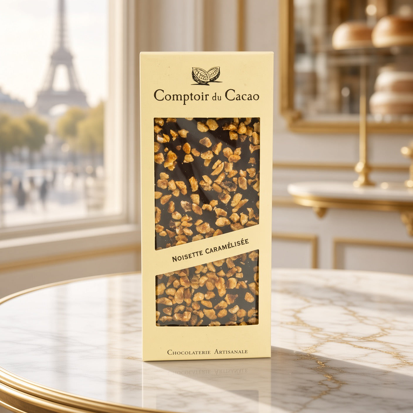 A box of Comptoir du Cacao Noisette CaramΓ©lisΓ©e chocolate, standing on a marble table with a blurred Parisian window and Eiffel Tower in the background.