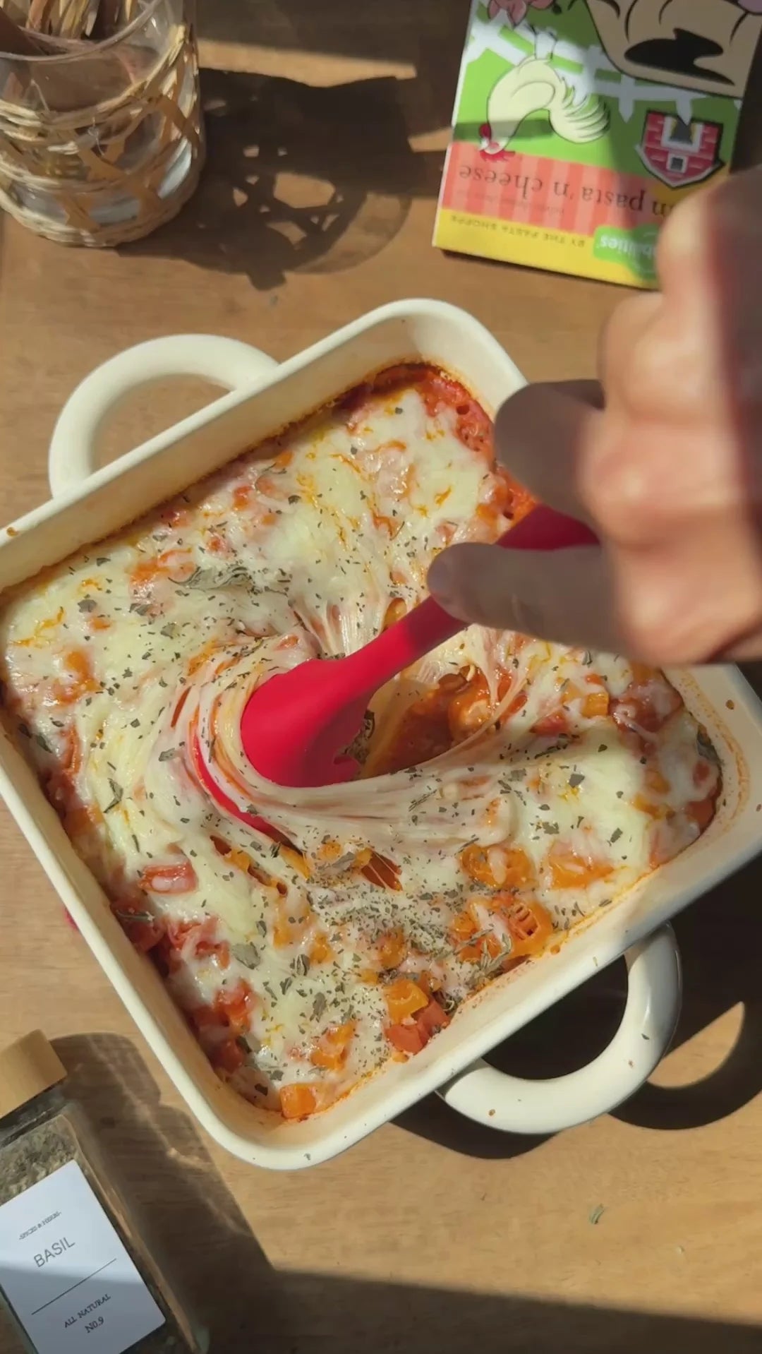 A close-up of a baked pasta dish in a white ceramic baking dish on a wooden table, with a hand holding a red spatula and a jar of seasoning nearby.