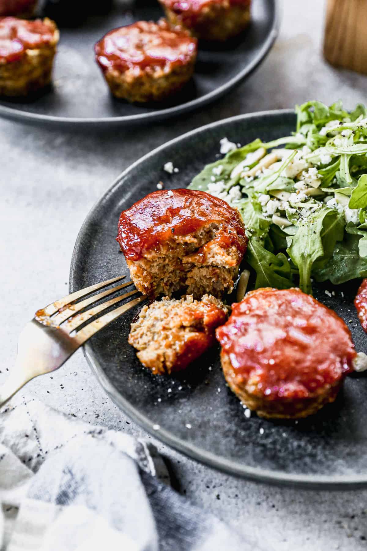 Two small meatloaf-style baked pieces topped with red sauce, served on a dark plate with a side of green salad and crumbled cheese, with a fork resting beside the food.