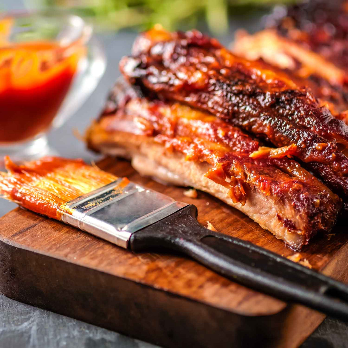 Sliced ribs on a wooden board with a brush and red sauce in the background
