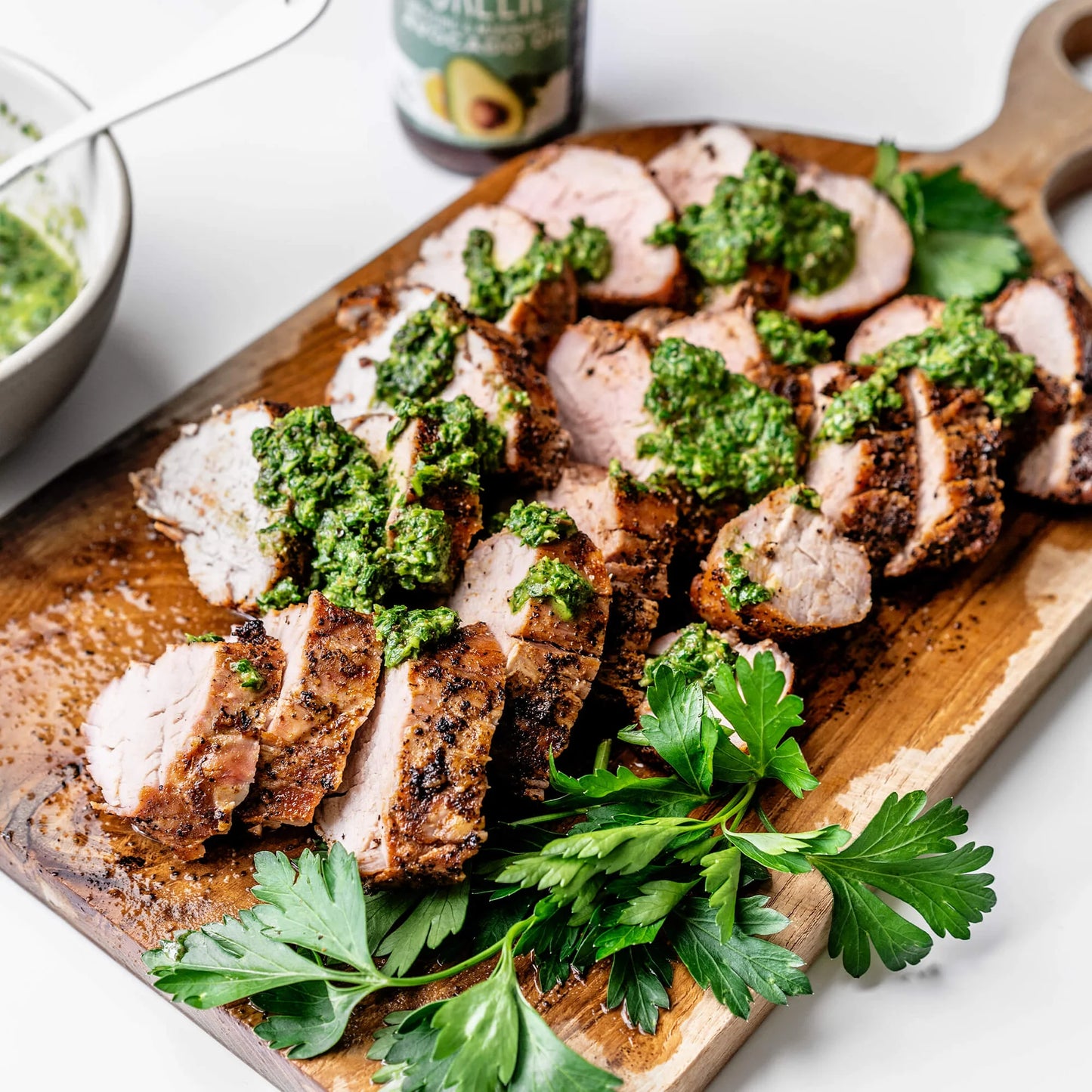 Sliced cooked meat topped with green herb sauce on a wooden serving board, garnished with parsley, with a bowl and bottle partially visible in the background.