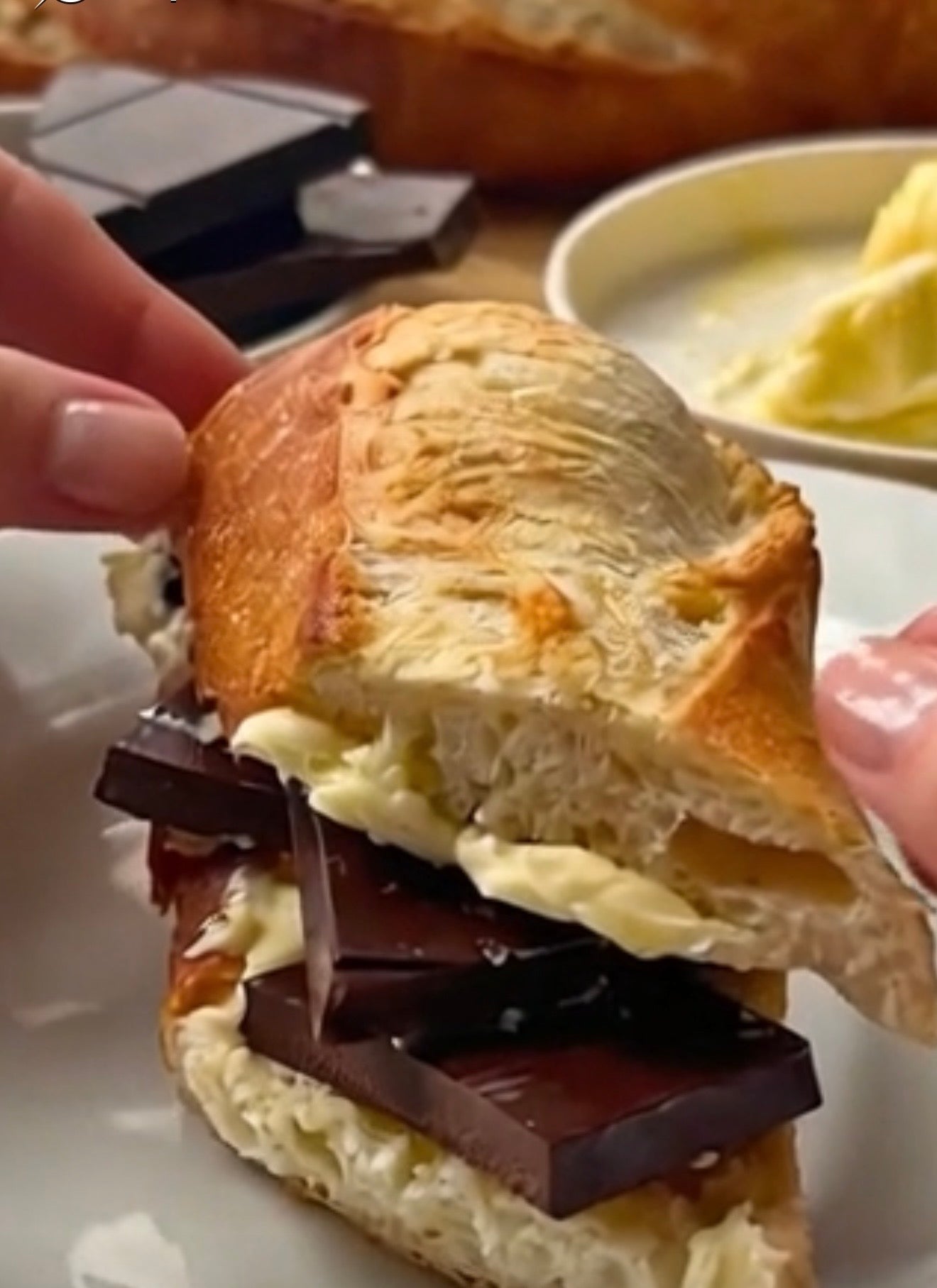 A close-up of a hand holding a half-eaten french baguette filled with melted buerre butter and dark chocolate pieces, with more chocolate and a butter dish blurred in the background.