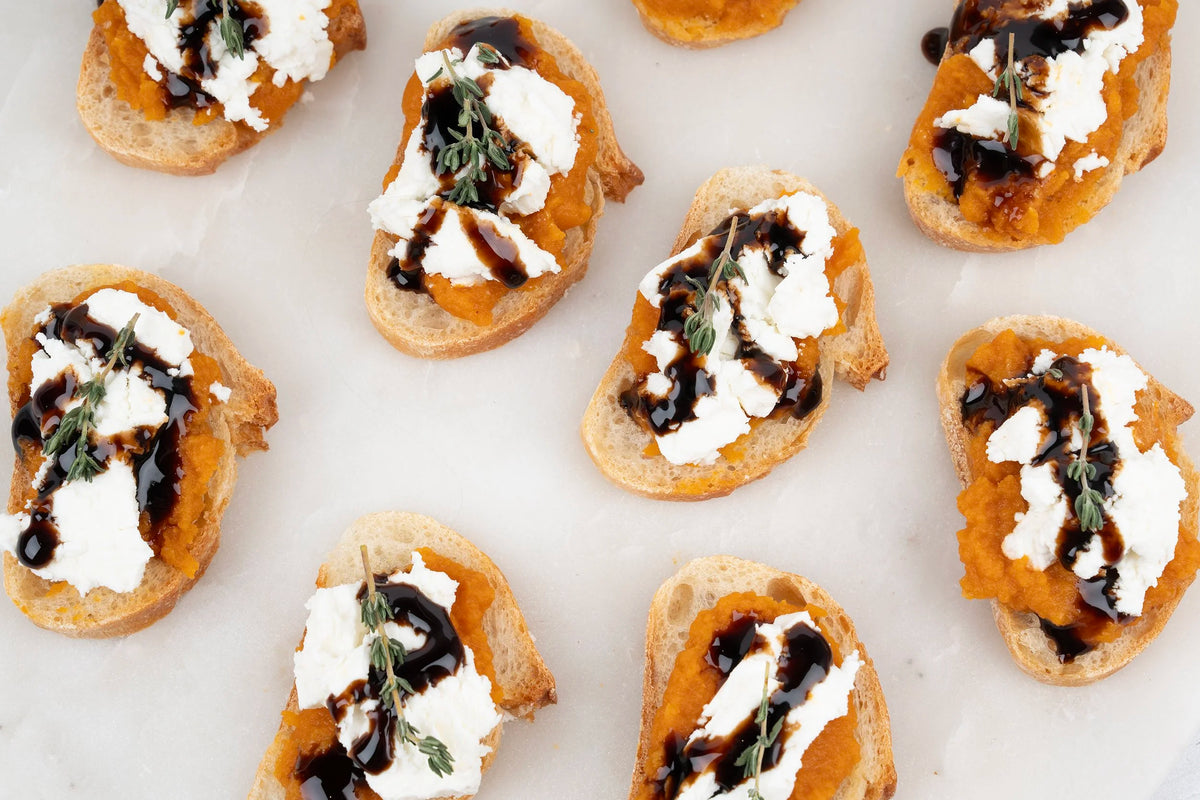 Multiple pieces of bruschetta arranged on a white marble surface, each topped with white cheese, orange pumpkin or sweet potato spread, dark balsamic drizzle, and a small herb sprig.