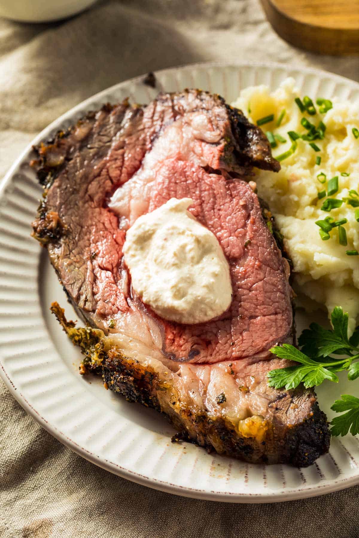 A close-up of a cooked prime rib roast served on a white plate with mashed potatoes and chives, topped with a dollop of white sauce, on a beige fabric surface.