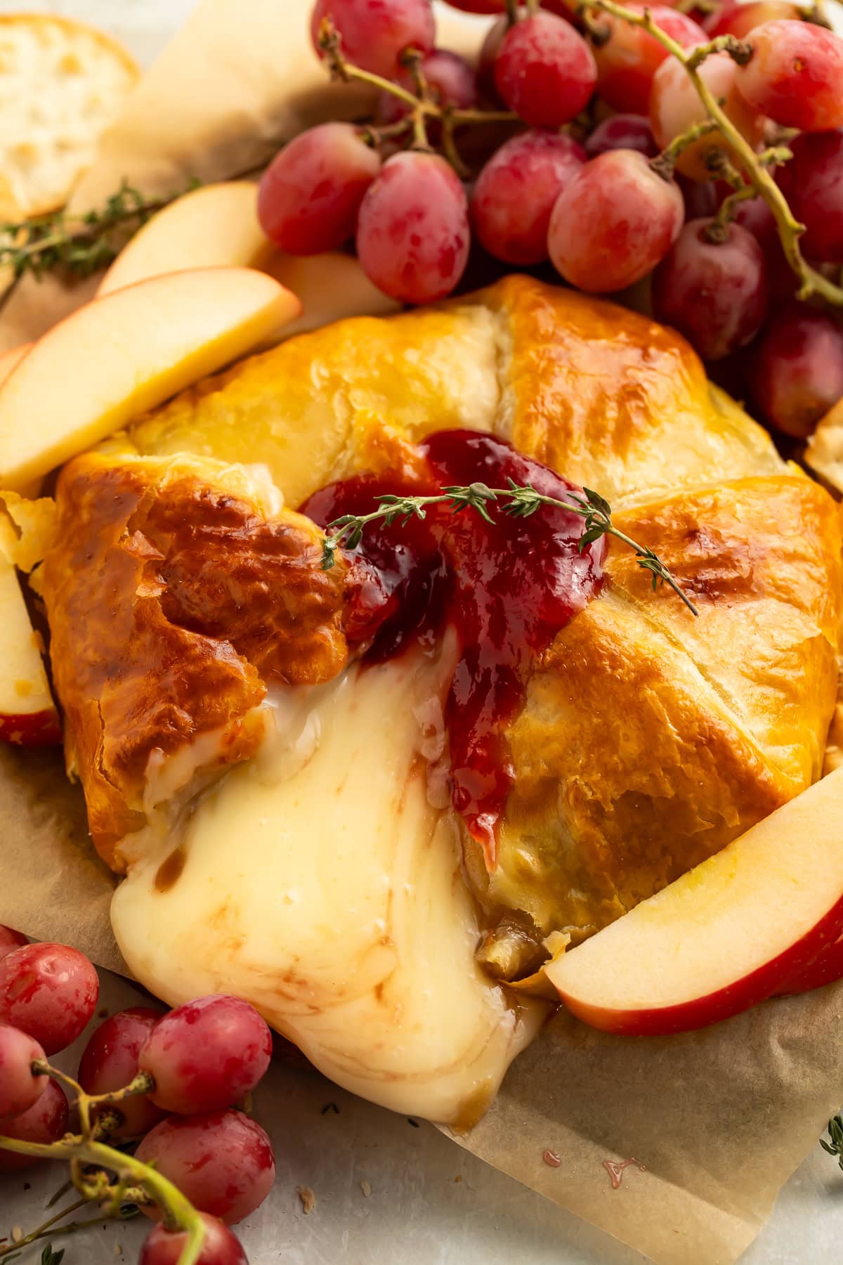 A close-up of a baked cheese pastry with a glossy red berry topping, melted cheese oozing out, surrounded by red grapes, apple slices, and more cheese on parchment paper.