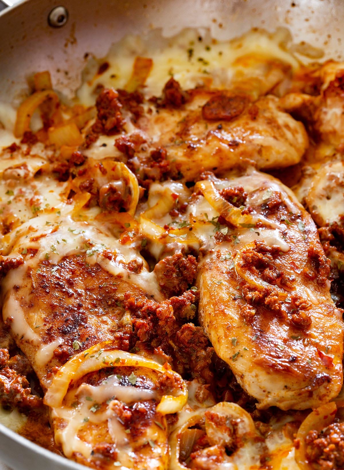 Close-up of baked chicken breasts topped with ground chicken chorizo melted cheese, browned breadcrumbs, and sliced onions in a skillet.
