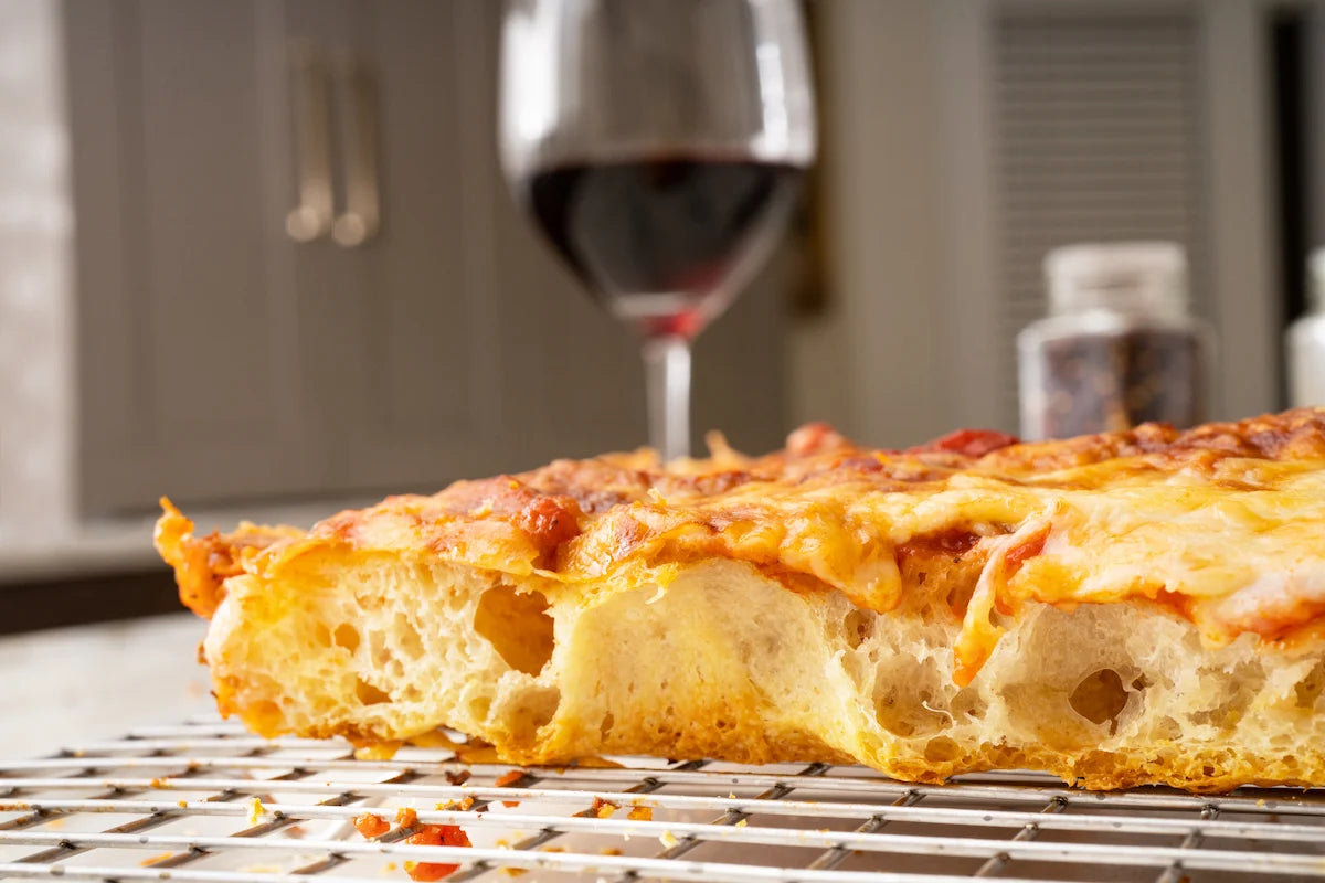 A close-up of a slice of cheesy pizza on a cooling rack, with a blurred glass of red wine in the background.