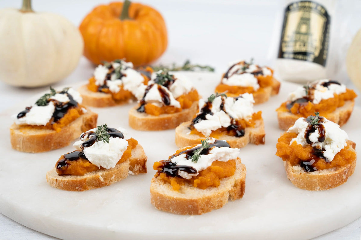 Small appetizer slices of toasted bread topped with mashed orange pumpkin, whipped cream, and dark balsamic drizzle, arranged on a white platter with pumpkins and a bottle in the background.