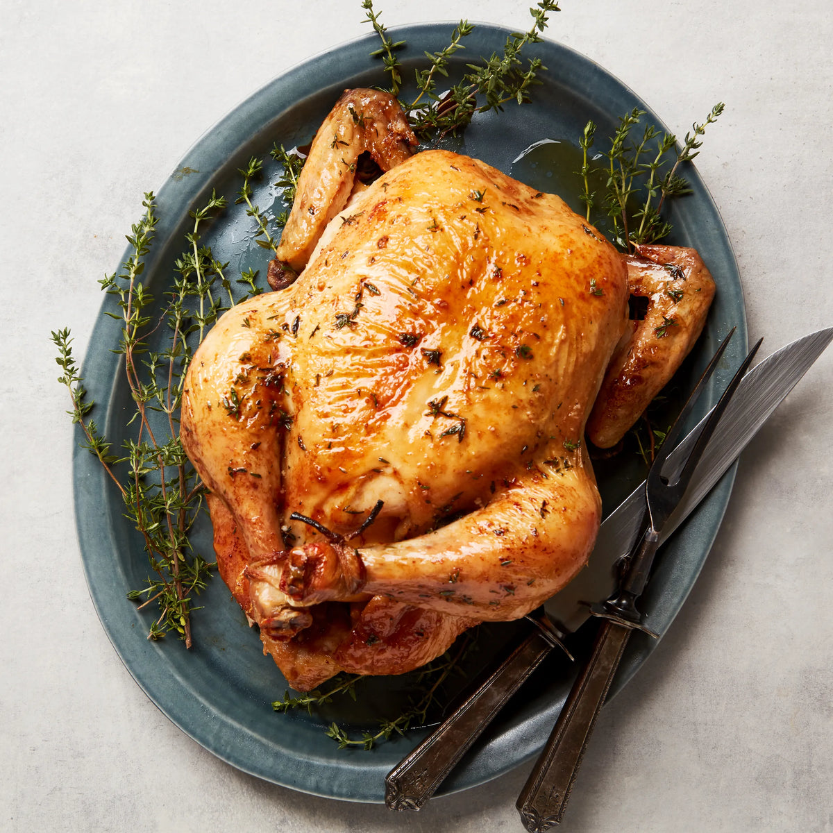 Roasted chicken on a blue plate with herbs and utensils on a light gray background