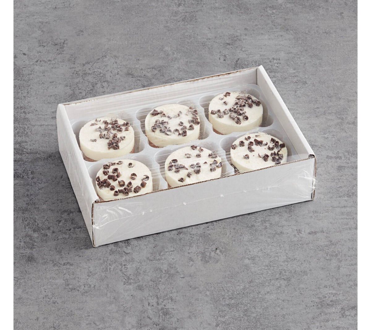 A white cardboard gift box containing six round white chocolate treats topped with dark chocolate chips, photographed on a gray textured surface.