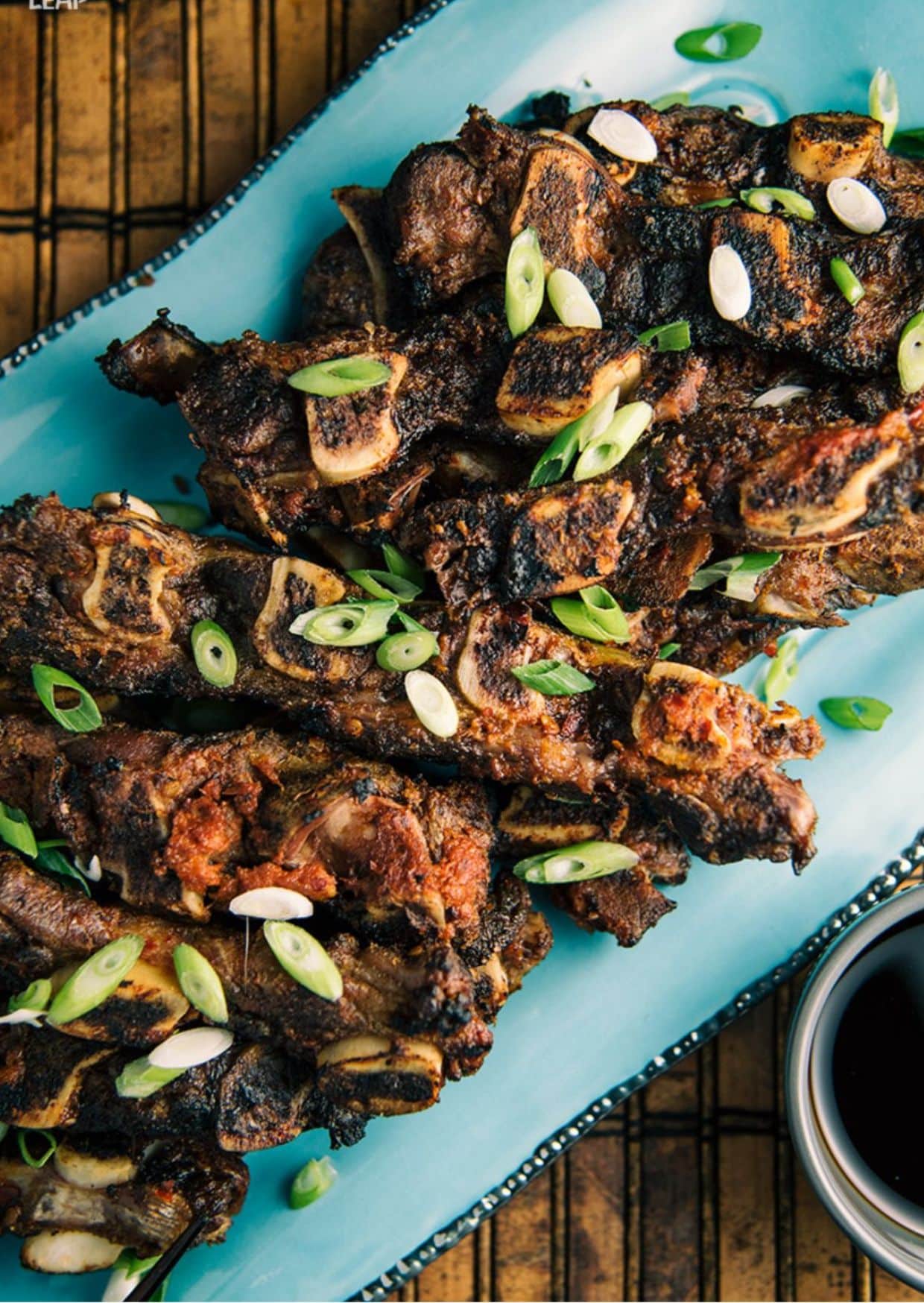Grilled short ribs garnished with sliced green onions on a light blue rectangular platter, set on a dark wooden table.