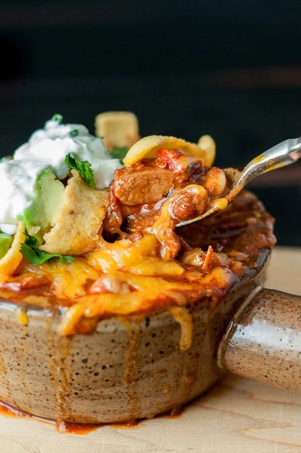 A close-up of a hearty chili with ground chicken chorizo bowl topped with melted cheese, sour cream, avocado, and tortilla chips, with a spoon lifting a portion of the chili.