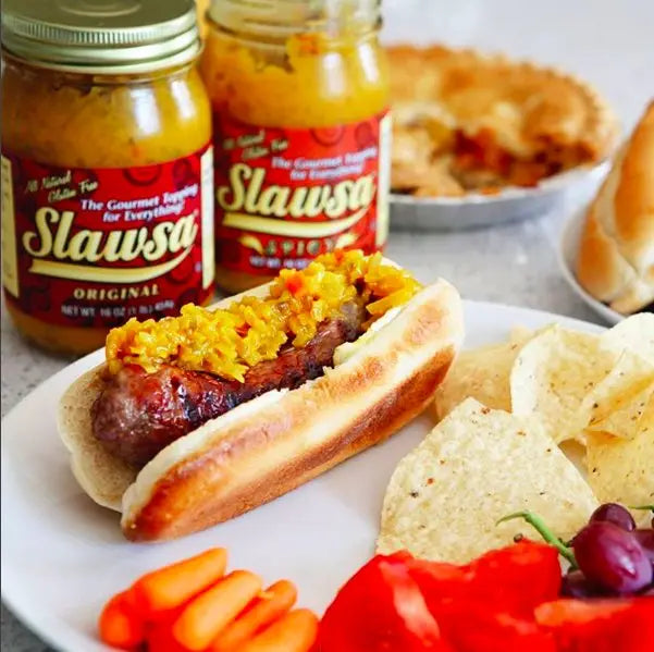 Hot dog with mustard on a bun, chips, carrots, and cherry tomatoes with Slawsa jars in the background.