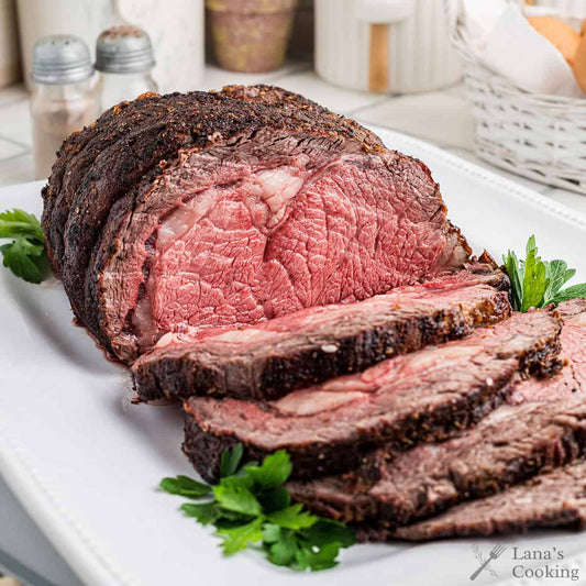 A sliced roast of prime rib cooked beef on a white platter, with green parsley garnish and blurred kitchen items in the background. 