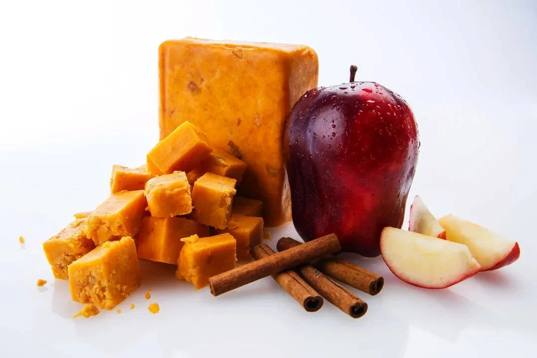 Block of cinnamon apple cheddar cheese with chunks, a red apple, and cinnamon sticks on a white background