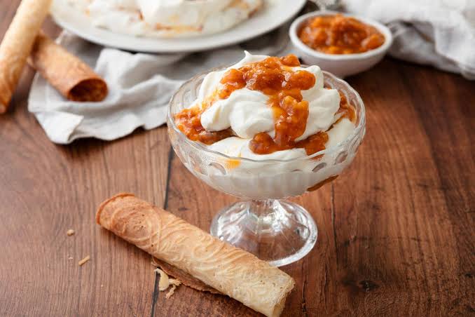 A dessert dish in a clear glass pedestal bowl topped with whipped cream and cloudberry fruit sauce, set on a wooden table with pastry pieces and a small bowl of sauce in the background.  