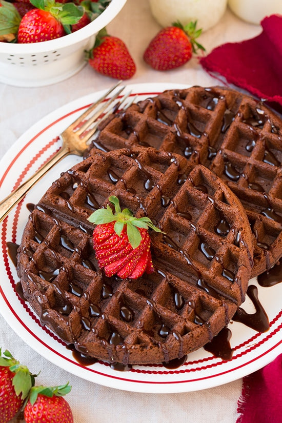 Two chocolate waffles topped with chocolate sauce and a fresh strawberry, served on a white plate with red trim. A gold fork rests on the plate, with additional strawberries and a white colander visible in the softly blurred background.