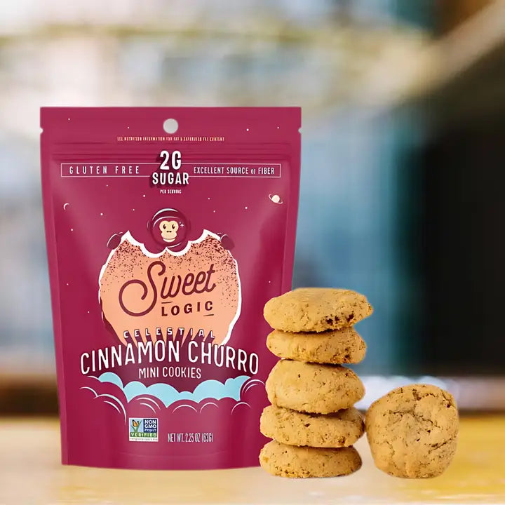 A bag of Sweet Logic Cinnamon Churro Mini Cookies stands on a light surface with several cookies stacked and placed beside the package. The bag is a deep pink/magenta color with white and peach graphics, and the background is softly blurred.