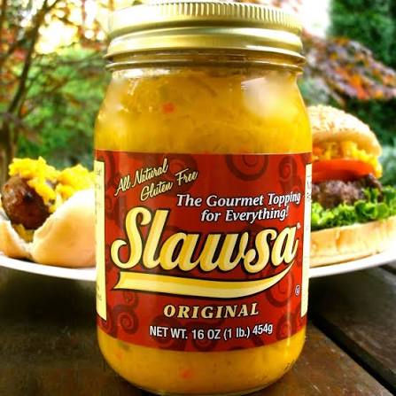 A jar of Slawsa Original gourmet topping, a yellow chunky relish in a clear glass jar with a gold lid and red label, set on a wooden table with burgers blurred in the background.