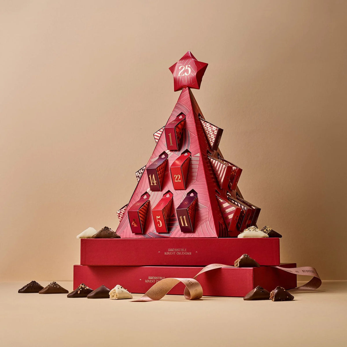 A red Christmas advent tree made of stacked gift boxes with numbered doors, displayed on matching red boxes with a ribbon and scattered chocolate pieces on a beige studio background.