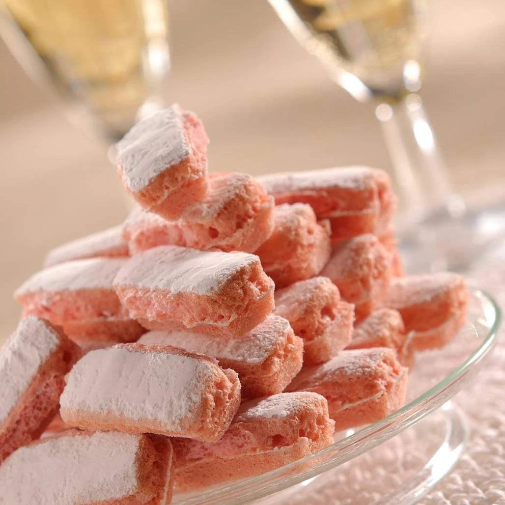 Pink ladyfinger cookies dusted with white powdered sugar, stacked on a clear glass plate with a blurred background of two champagne glasses.