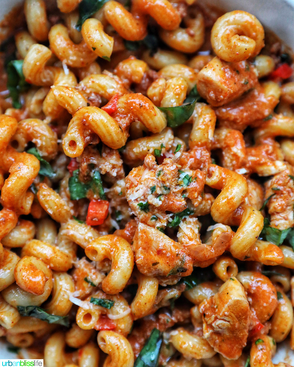 A close-up overhead shot of a cooked pasta dish in a pan. The dish features rotini pasta coated in a rich, orange-red tomato sauce, mixed with vground chicken chorizo, chopped green herbs, and grated cheese.