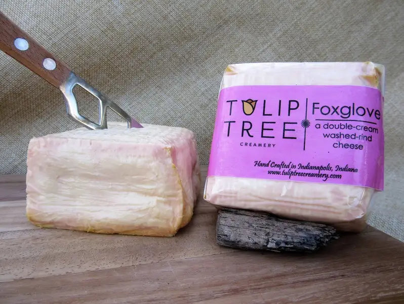 A block of Tulip Tree Creamery Foxglove washed and aged cheese stands next to its paper-wrapped package on a wooden surface against a burlap backdrop, with a cheese knife inserted into the cheese.