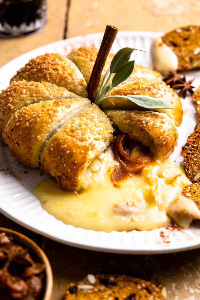 A baked pumpkin-shaped bread roll topped with melted cheese, a cinnamon stick, and green leaves, served on a white plate with toasted bread slices on a rustic wooden table.