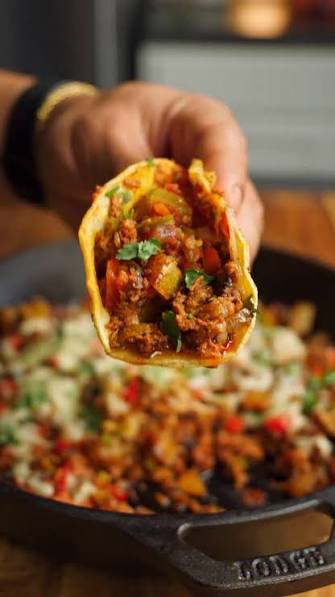 A hand holds a stuffed taco filled with seasoned ground chicken chorizo, diced vegetables, and cilantro above a cast iron skillet filled with the same taco filling, with a blurred kitchen background.