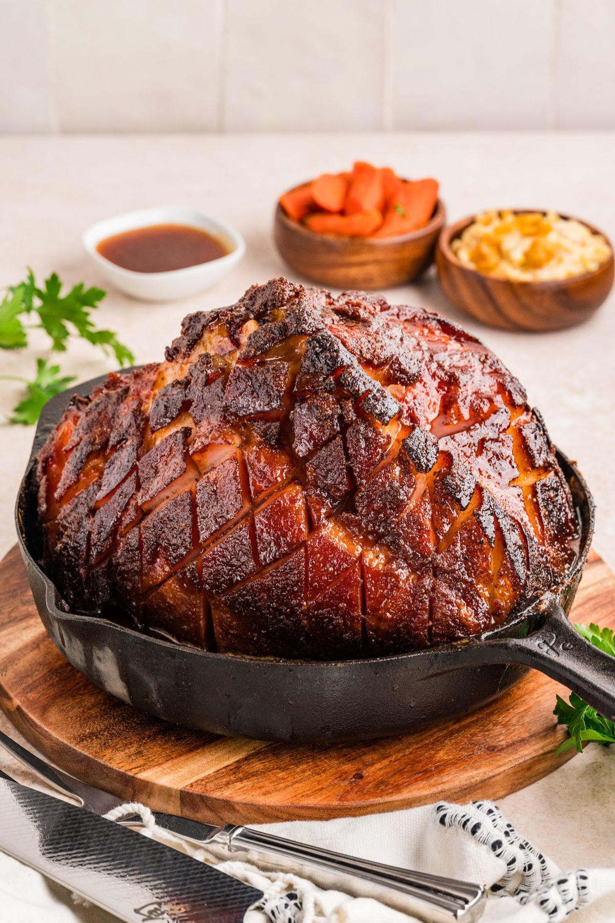 A roasted ham with a golden-brown glazed crust and deep scoring, served in a black cast-iron skillet on a round wooden board. Small bowls of dipping sauce and carrot sticks are blurred in the background.