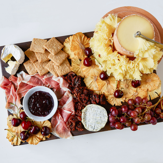 An overhead flat-lay of a charcuterie and cheese board on a white background, featuring assorted cheeses, crackers, cured meats, dried fruit, grapes, and a small bowl of dark preserves.