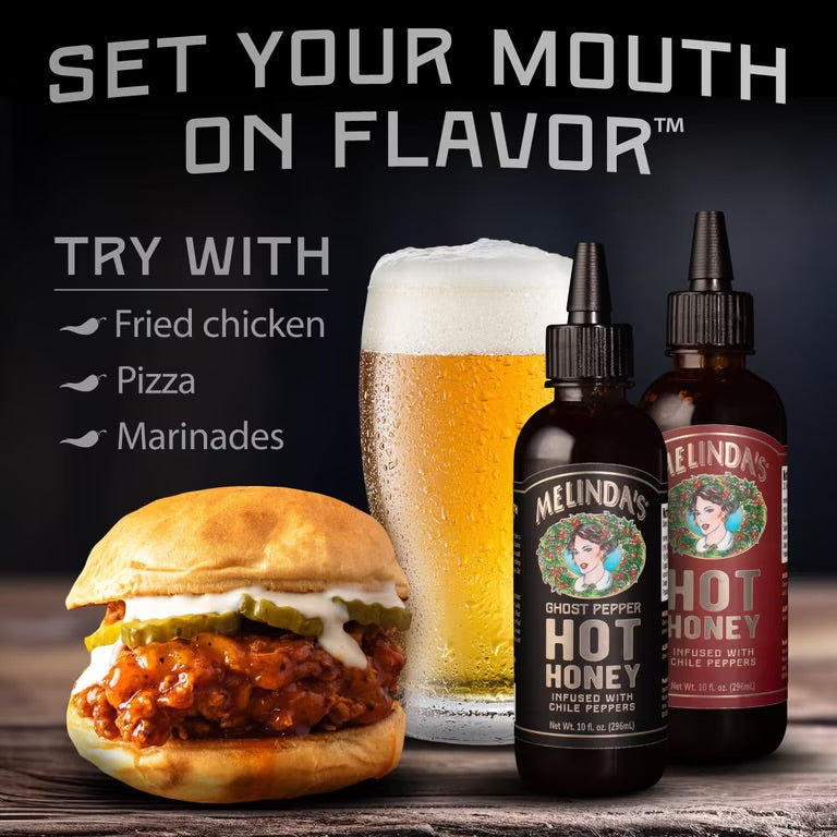 Melinda’s Classic and Ghost Pepper Hot Honey bottles beside a chicken sandwich and a glass of beer
