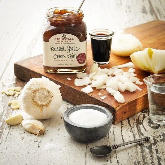 Stonewall Kitchen roasted garlic onion jam on a wooden cutting board with onions, garlic, and salt.