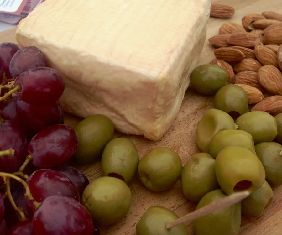 A close-up of a Foxglove cheese board featuring a large block of cheese, green olives, red grapes, almonds, and a wooden toothpick on a wooden cutting board.