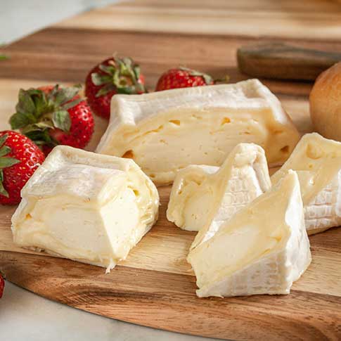 Soft Trillium cheese wedges and a larger rectangular cut on a wooden cutting board, styled with fresh red strawberries and a round bread or pastry in the background.