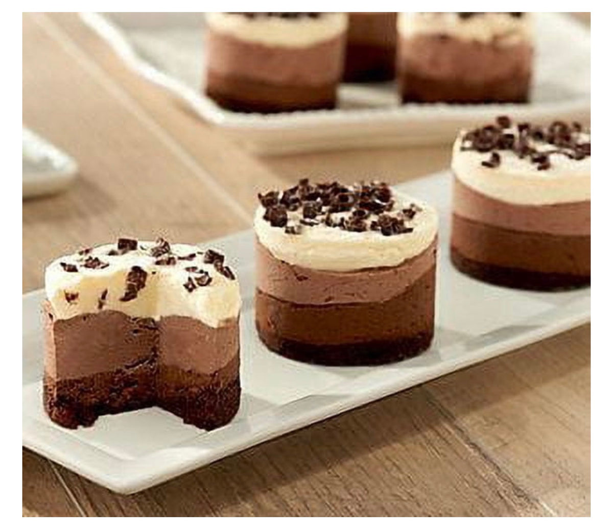 Three chocolate layer desserts topped with cream and chocolate crumbs, arranged on a white rectangular platter on a wooden table.