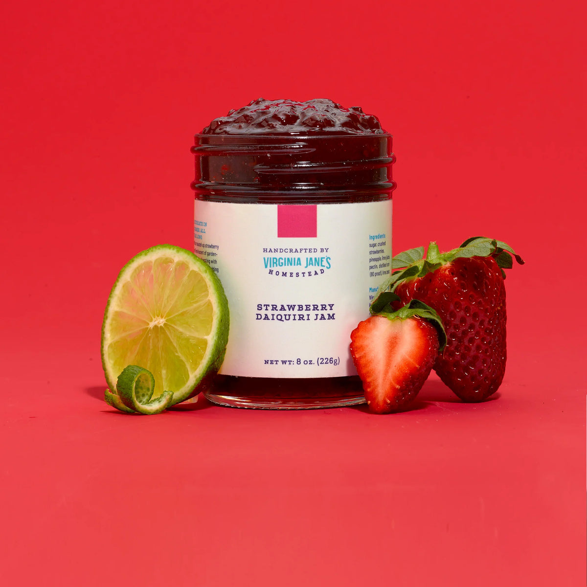 A jar of Virginia Jane's Romster Strawberry Daiquiri Jam is displayed on a vibrant red background, styled with fresh ingredients including whole strawberries, a sliced lime, and a lime peel.