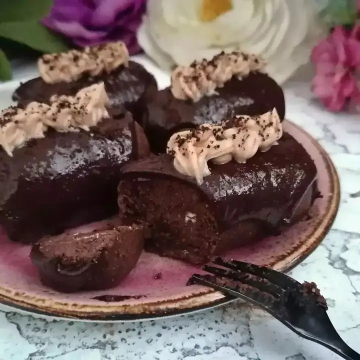 Chocolate cake rolls with piped cream frosting and cocoa powder sprinkles, served on a pink plate with a fork, set against a blurred floral background.