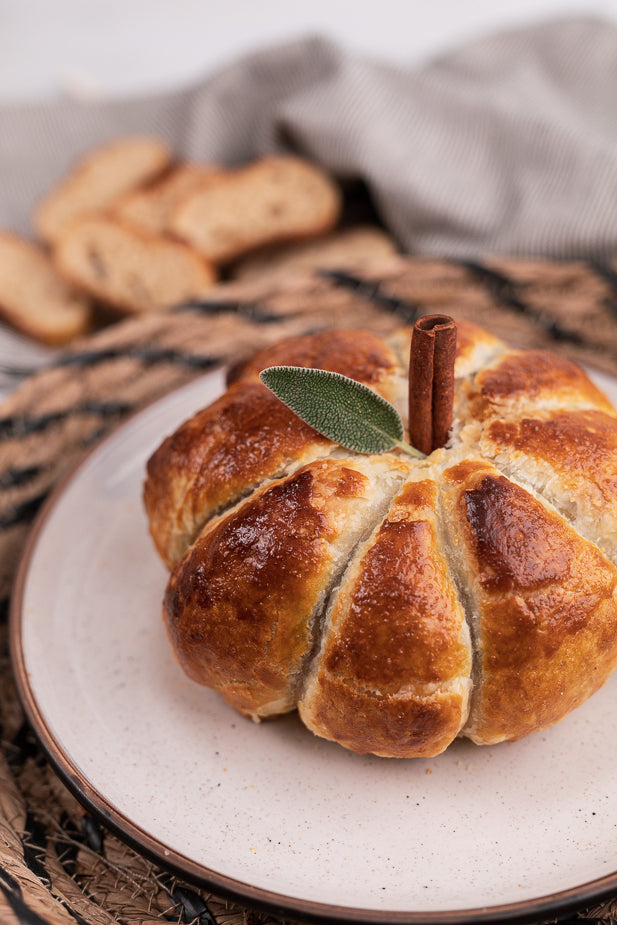 A pumpkin-shaped bread roll with golden-brown baked sections, a cinnamon stick stem, and a green leaf garnish, served on a light beige ceramic plate.