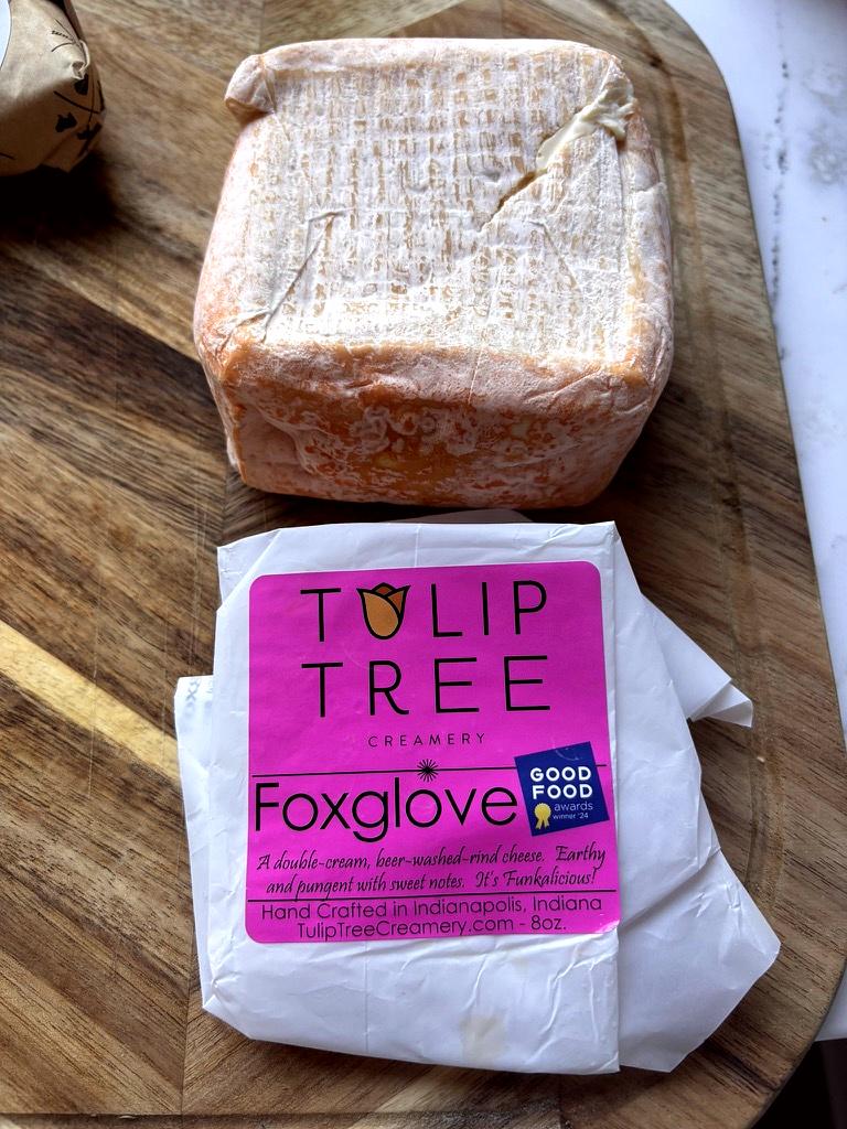 A block of Tulip Tree Creamery Foxglove cheese wrapped in white paper with a bright pink label, placed on a wooden cutting board below a second wrapped cheese block.
