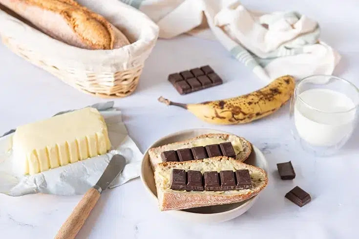A styled food scene on a light marble surface showing a basket of baguettes, a block of butter, a glass of milk, and two slices of bread topped with dark chocolate squares. A banana and a cloth are also visible.