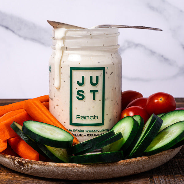 A jar of Just Ranch dressing sits on a wooden board with sliced cucumbers, carrot sticks, and cherry tomatoes, set against a white marble background.