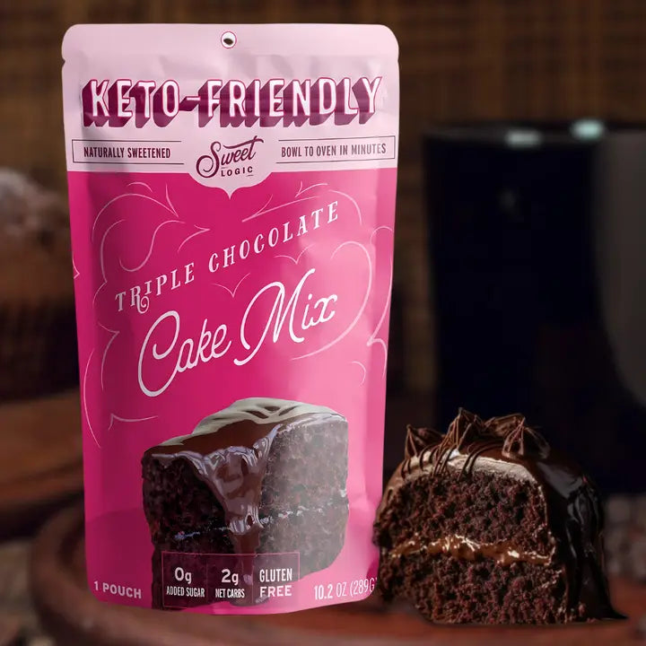 A pink stand-up pouch of Sweet Logic Keto-Friendly Triple Chocolate Cake Mix stands on a wooden surface next to a sliced chocolate cake, with a blurred kitchen background.