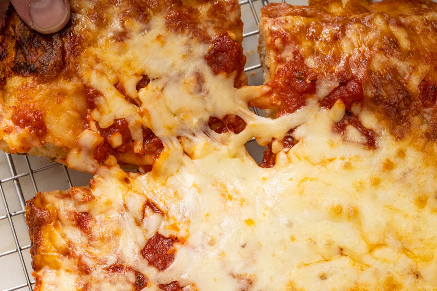 Close-up of a cheesy baked pizza with tomato sauce, showing melted white cheese stretching between slices on a cooling rack.